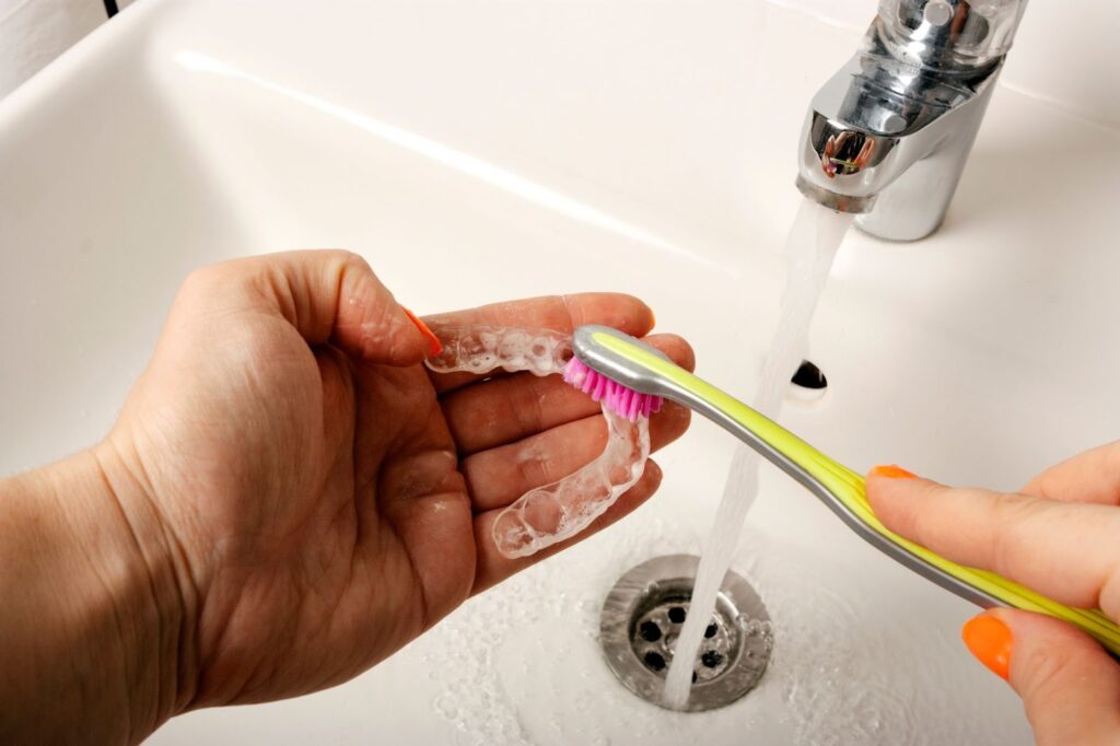 Hand holding a clear retainer under running water, brushing with a soft toothbrush, emphasizing retainer care for orthodontic maintenance at TopSmiles Pediatric Dentistry & Orthodontics in Winnipeg.