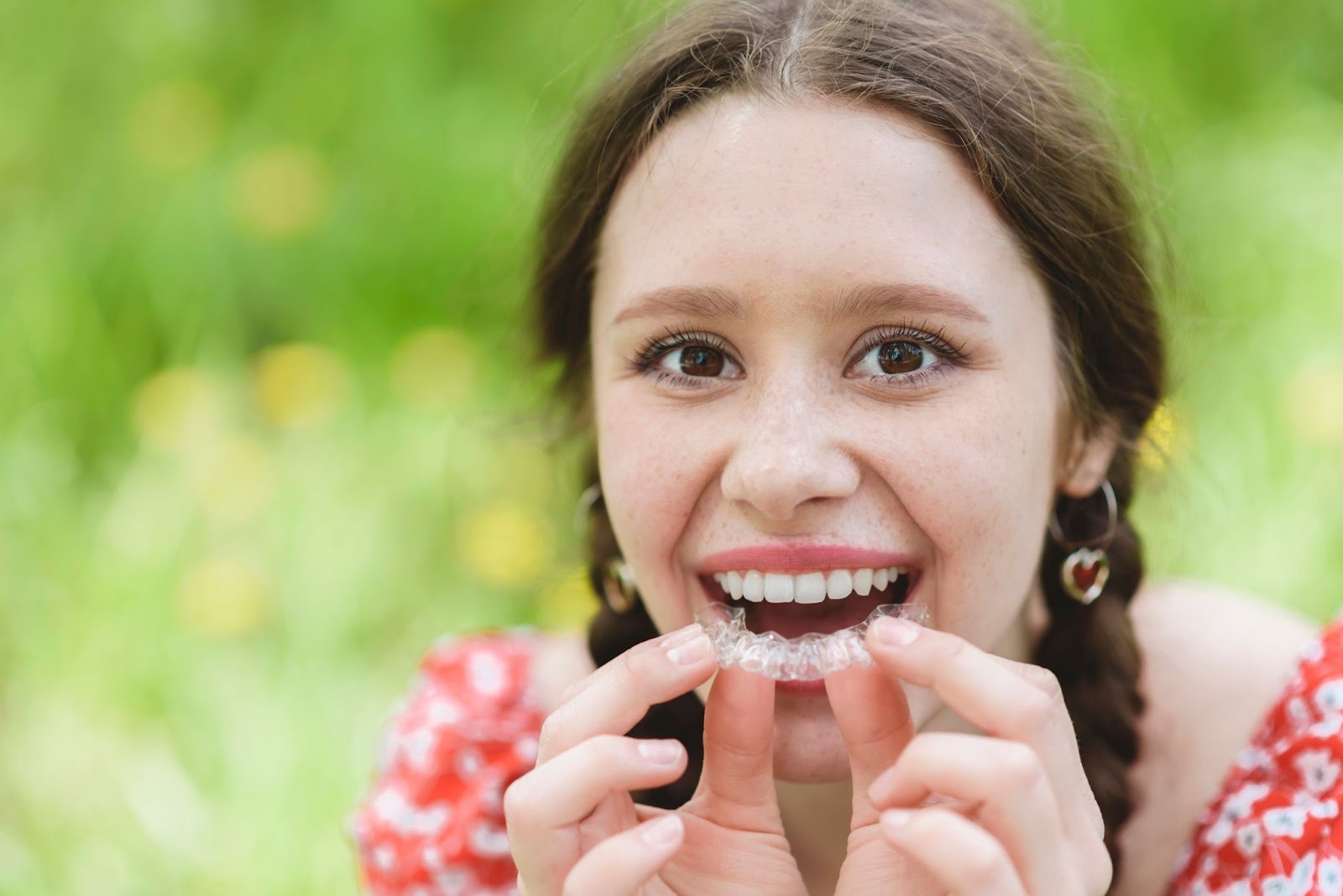 Young woman smiling while holding a clear retainer, showcasing dental alignment post-braces, with a blurred green background, emphasizing orthodontic care and retainers at TopSmiles Pediatric Dentistry & Orthodontics in Winnipeg.